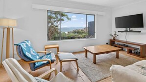 A modern living room with a large window, a flat-screen TV, and a wooden coffee table. - Kareelah | Ocean Views & Walk to Beach (Guerilla Bay)