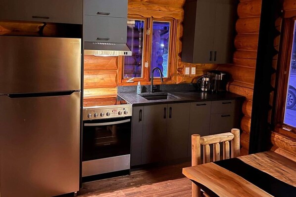 A compact kitchen with a stainless steel refrigerator, a stove, and a sink. There is a wooden dining table and a chair. The kitchen features wooden walls and a window with a view outside.