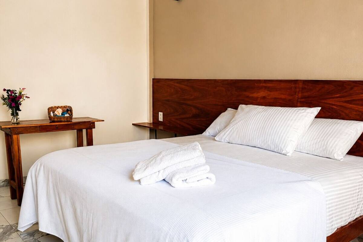 A neatly made bed with white linens and towels, a wooden headboard, a small bedside table with a vase and basket, and marble-patterned flooring.