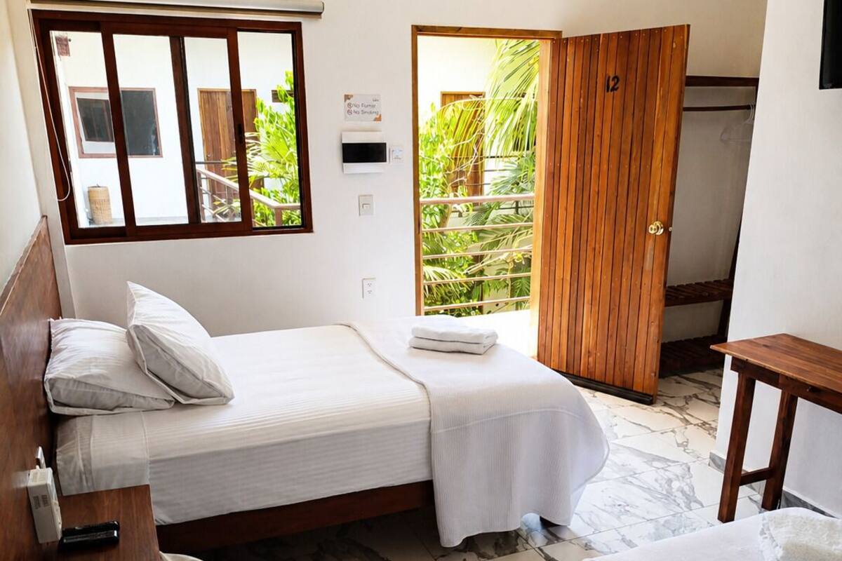 A room with two beds, a wooden door, a small table, and a window with a view of greenery.