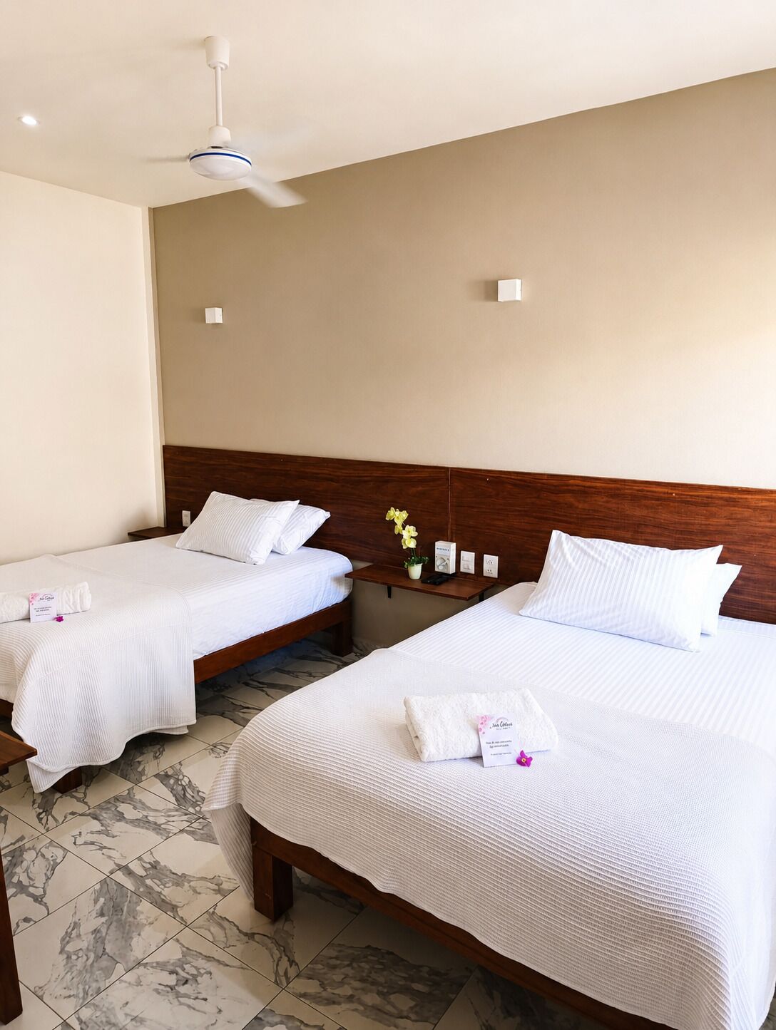 Habitación de hotel con dos camas, cabecera de madera, suelo de mármol, ventilador de techo y una mesita de noche con un florero.