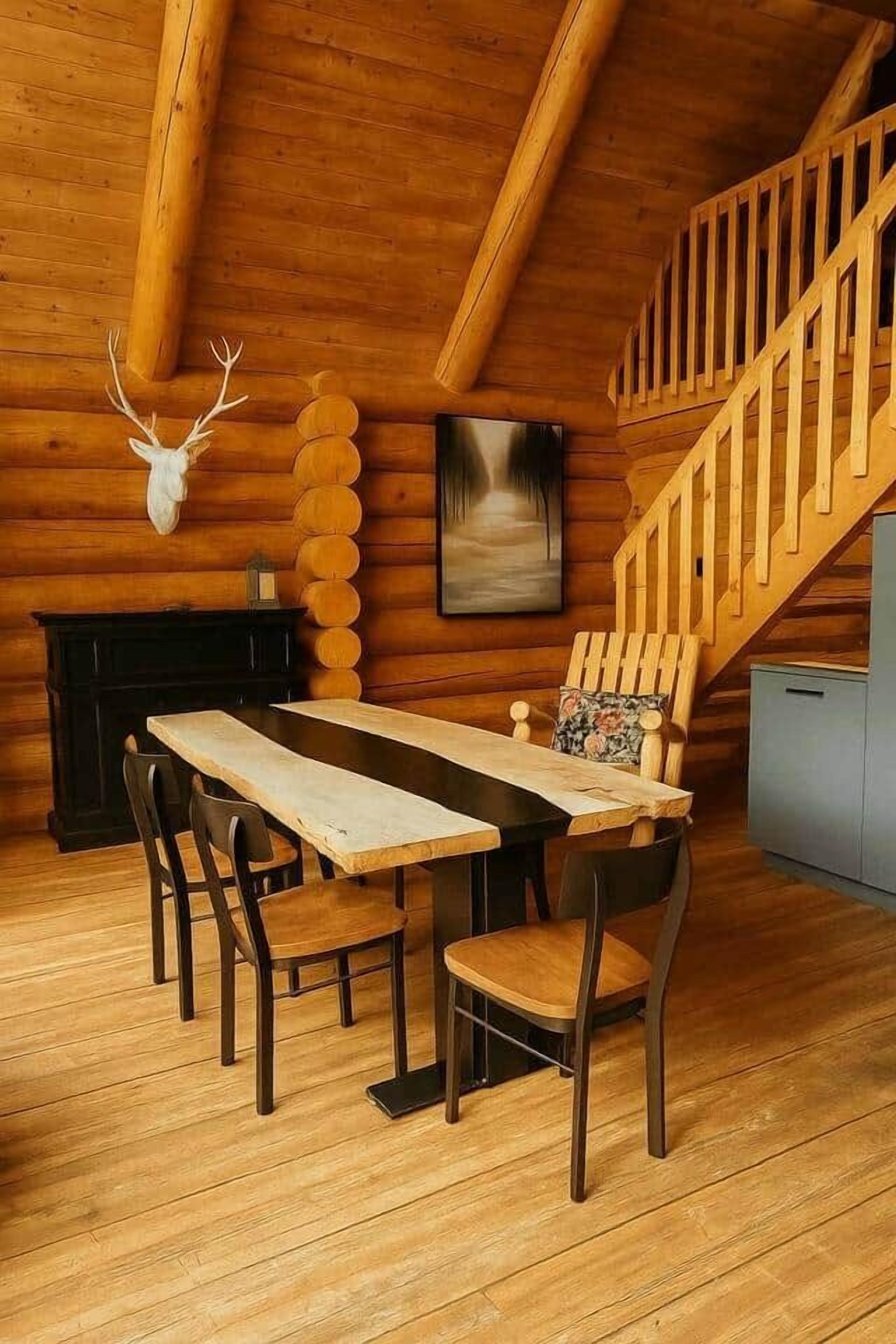 A rustic log cabin interior with a wooden dining table, four chairs, a fireplace, a deer head mounted on the wall, and a staircase.