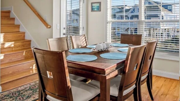 A dining area with a wooden table and chairs, a staircase, and large windows with blinds. - "Tailwinds and Tides" Ocean and Marsh Views on Holden Beach NC (Holden Beach)