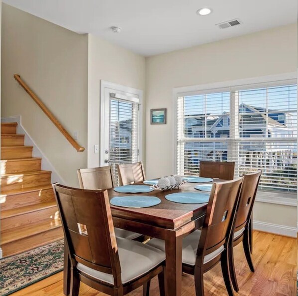 A dining area with a wooden table and chairs, a staircase, and large windows with blinds. - "Tailwinds and Tides" Ocean and Marsh Views on Holden Beach NC (Holden Beach)