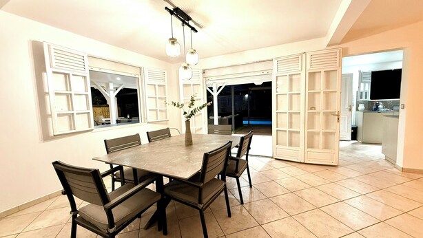 A dining area with a large table, chairs, and a vase with plants. There are windows with shutters and a view of the outside.