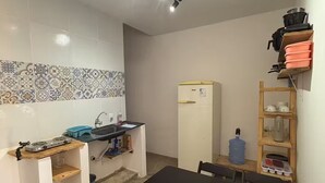 A compact kitchen with a sink, stove, and a water dispenser. There is a dining table with chairs and a shelf with kitchenware. - House on the beach street in Gostoso (Touros)