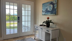 Een kamer met een witte deur, een klein wit bureau met een bloevas, en een schilderij van een ruige zee aan de muur.