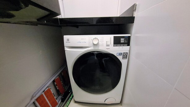 Une petite machine à laver blanche, dotée d’un panneau de commande, située dans un petit espace buanderie aux murs carrelés et où une prise électrique est visible.