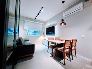 A modern living room with a sofa, dining table, chairs, a TV, and a window with blinds. - Leblon- Design e aconchego.  (Rio de Janeiro)
