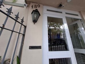 A building facade with a numbered address, a metal gate, a window with a view, and a wall-mounted light fixture. - Leblon- Design e aconchego.  (Rio de Janeiro)