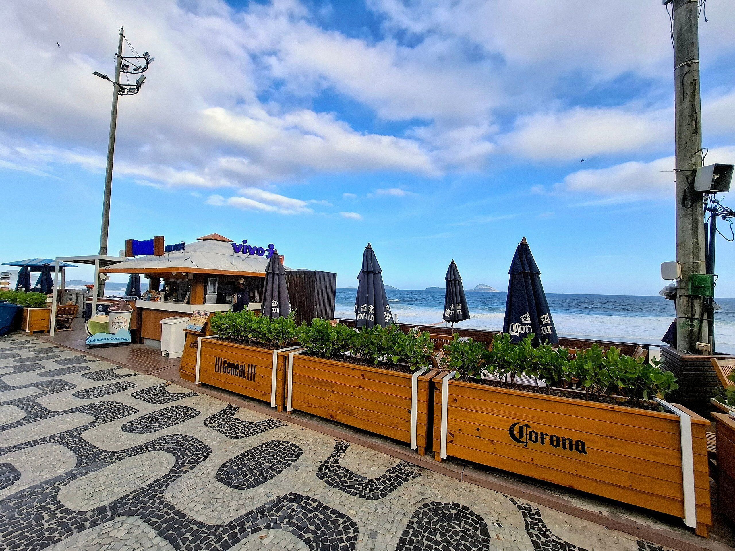 Restaurante à beira-mar com mesas ao ar livre, vista para o oceano e jardineiras de madeira com o logotipo Corona.