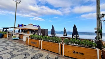 Restaurante à beira-mar com mesas ao ar livre, vista para o oceano e jardineiras de madeira com o logotipo Corona.