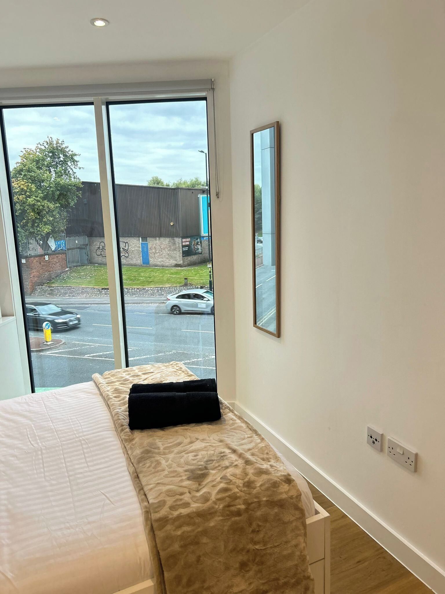 A bedroom with a bed, a window with a view of the street, and a wall-mounted mirror.