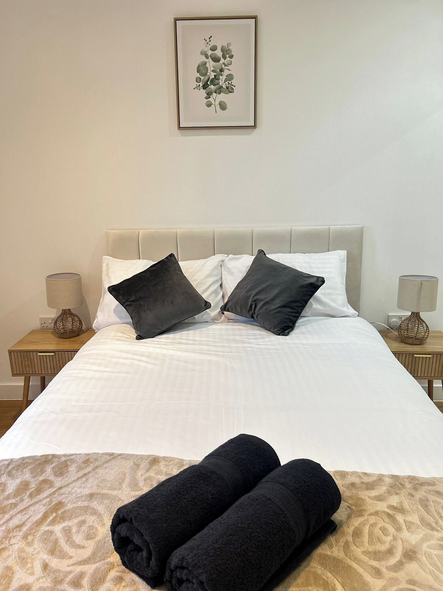 A neatly made bed with white linens and two black pillows, a framed botanical print on the wall, and two rolled towels on the bed.