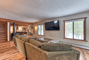 A spacious living room with a large sectional sofa, a flat-screen TV mounted on the wall, and large windows offering a view of the outdoors. - Lakehouse Sleeps 11, Golf, Trails, Lake, Fireplace, True 4-Season (St. Helen)