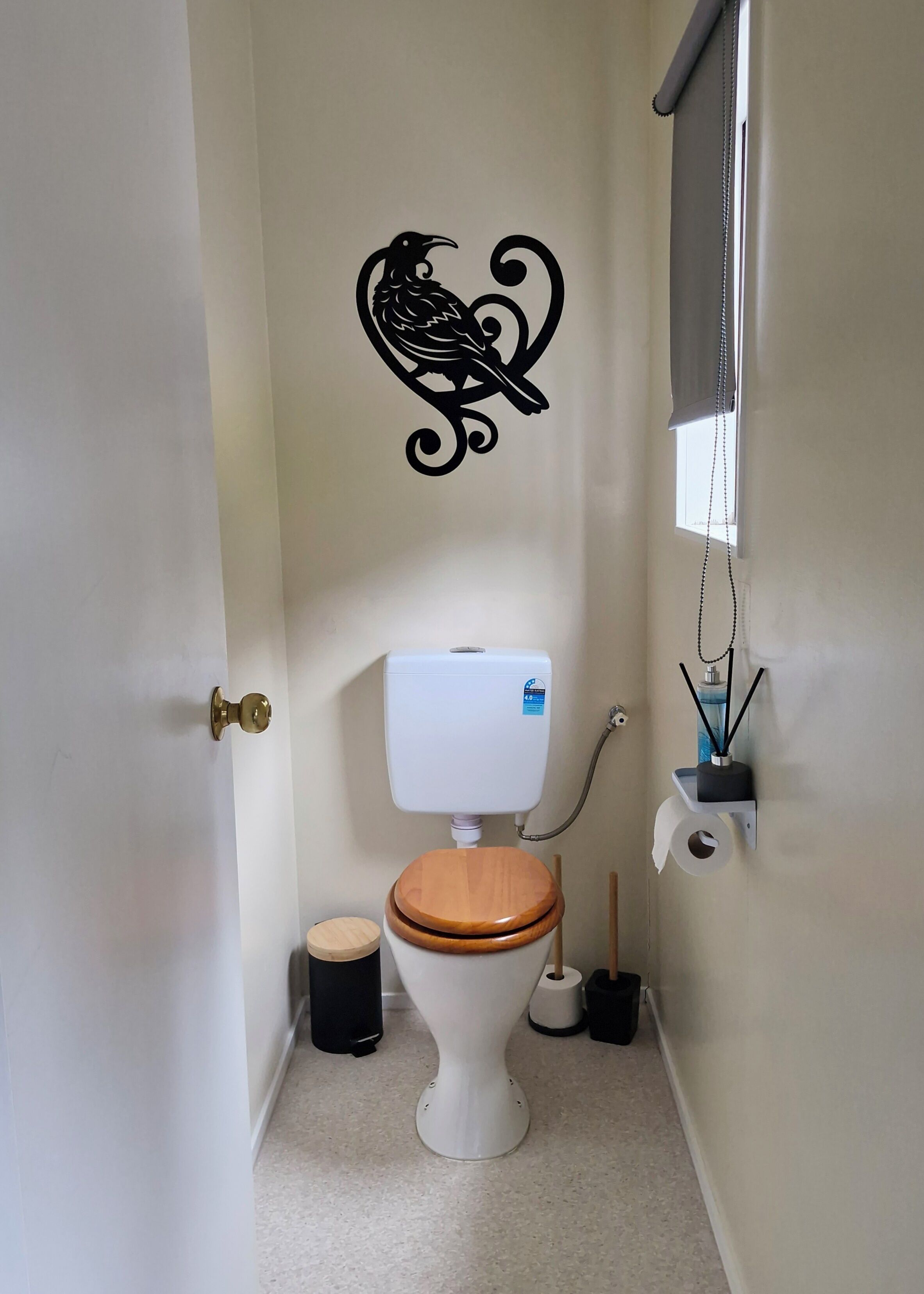 Een kleine badkamer met toilet, een houten deur, een zwarte muursticker van een vogel, een raam met zonwering en een toiletrolhouder met een rol.