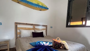 Ett sovrum med en säng, en sänggavel i trä, en väggdekoration med strandtema, ett fönster och en flätad korg på sängen.
