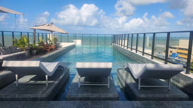 A rooftop infinity pool with lounge chairs and a glass railing overlooking the ocean.