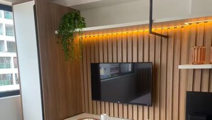 A compact living space with a dining area, a flat-screen TV mounted on a wooden wall, a small kitchen area, and a bed with a green cover.