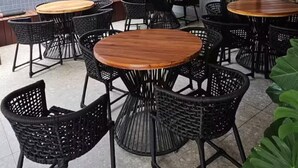 An outdoor patio area with round wooden tables and black wicker chairs, featuring a tiled wall and a glass door leading to the interior.
