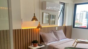 A modern bedroom with a bed, bedside table, hanging pendant lights, a small plant, and a window with a city view.