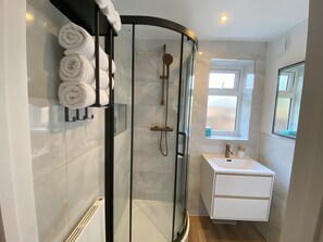 Une salle de bain moderne avec une douche à parois de verre, des carreaux blancs, un lavabo blanc avec un miroir et une fenêtre laissant entrer la lumière naturelle.