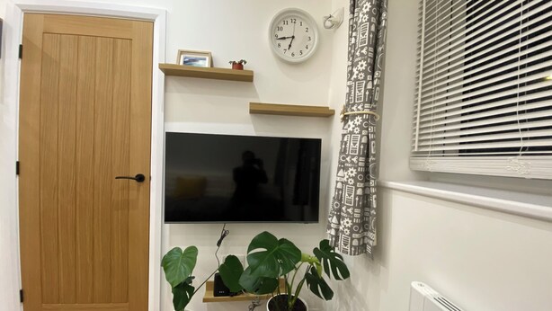 Een kamer met een houten deur, een wandtelevisie, twee potplanten, een klok en kamergordijnen met een patroon.