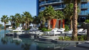 A pool area with lounge chairs and palm trees, surrounded by a modern building with multiple balconies. - Cozy 1-bedroom apartment in Business Bay, Downtown (Dubai)