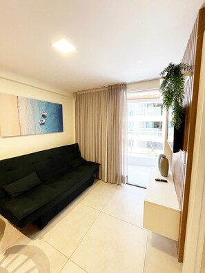 A modern living room with a dark green sofa, a painting of a beach scene, a window with curtains, and a plant on a wooden shelf. - Puerto Ventura By Day by Day (João Pessoa)