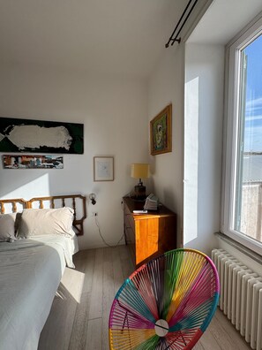 A bedroom with a bed, a dresser, a lamp, a colorful fan, and a window with a view. - The Painter's House—The Muse's Apartment (Roma)