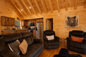 A cozy cabin interior with a wooden interior, a sofa, two recliners, a refrigerator, and a bear painting on the wall. - Romantic Log Cabin • Hot Tub + Vinyl Records • 15 Min to Ellijay (Ellijay)