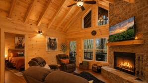 A cozy cabin interior with a fireplace, wooden walls, a ceiling fan, and a large window overlooking a forest. - Romantic Log Cabin • Hot Tub + Vinyl Records • 15 Min to Ellijay (Ellijay)