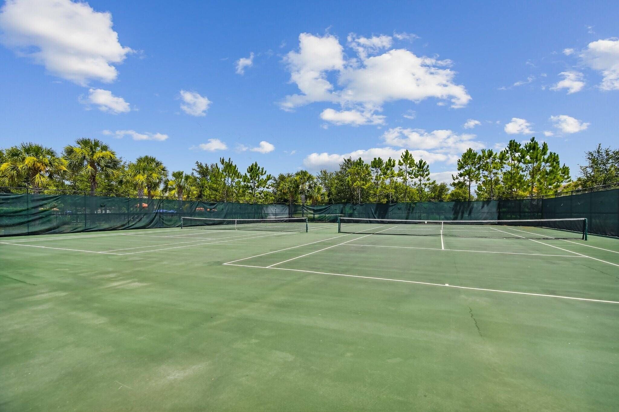 An outdoor tennis court with a net, surrounded by trees and a clear sky.