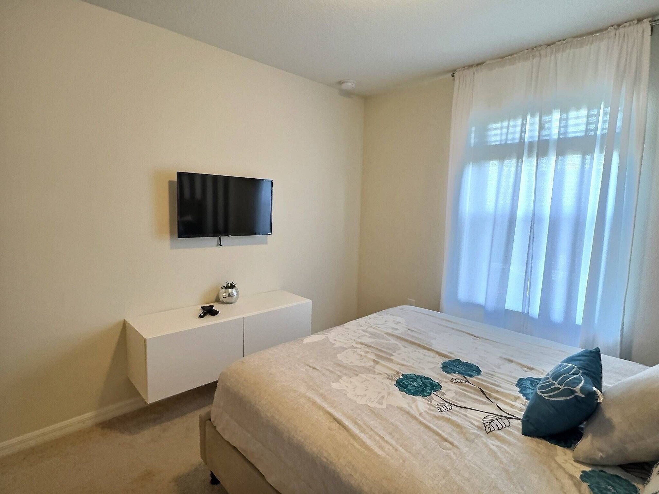 A bedroom with a bed, a TV mounted on the wall, a bedside table, and a window with curtains.
