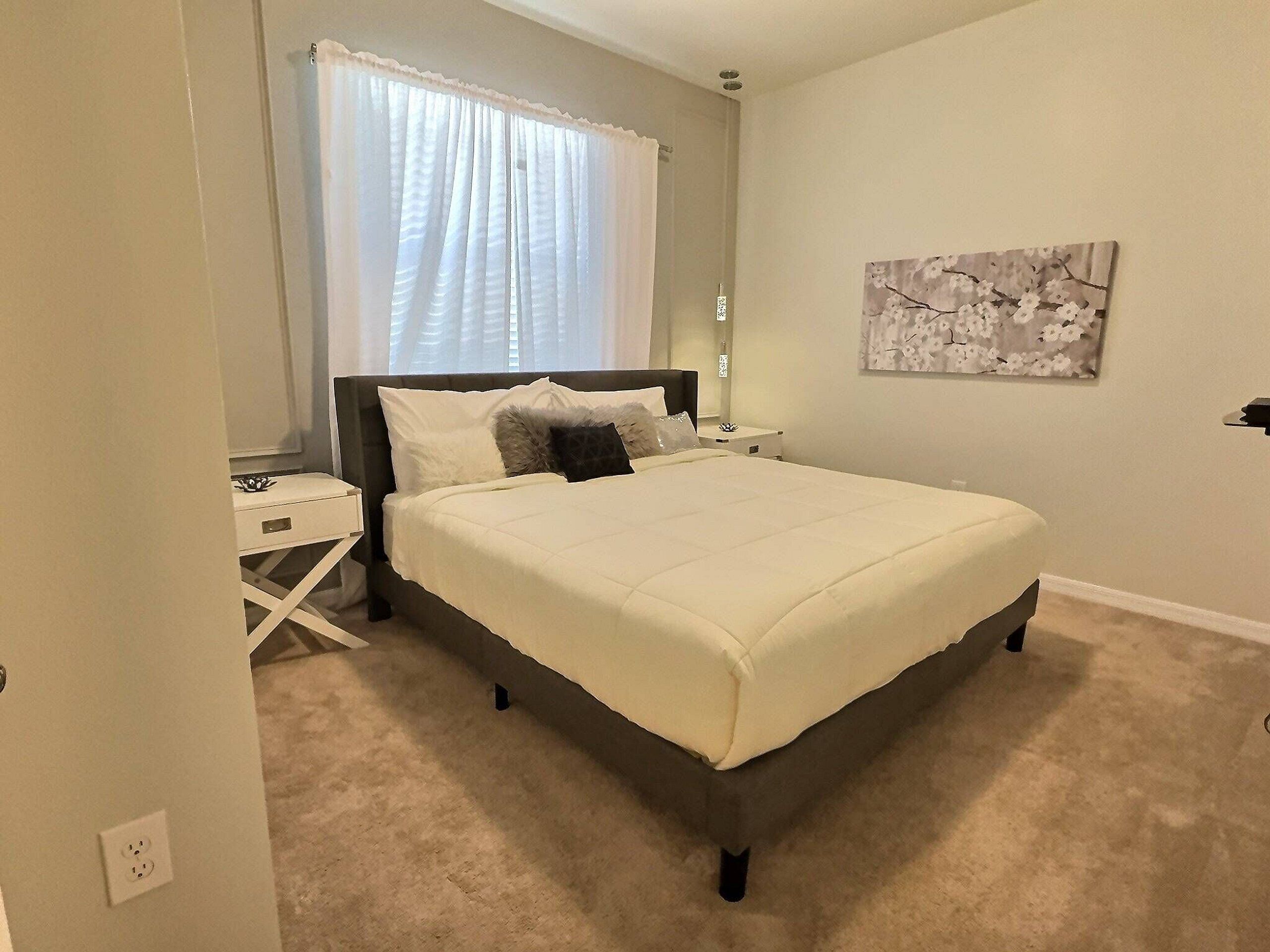 Un dormitorio con cama, mesitas de noche, una obra de arte en la pared y una ventana con cortinas.