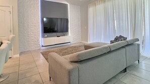 A modern living room with a flat-screen TV mounted on a white wall, a grey sofa, and a large window with white curtains. - Solterra 5BR Villa - Private Pool & Resort (Davenport)