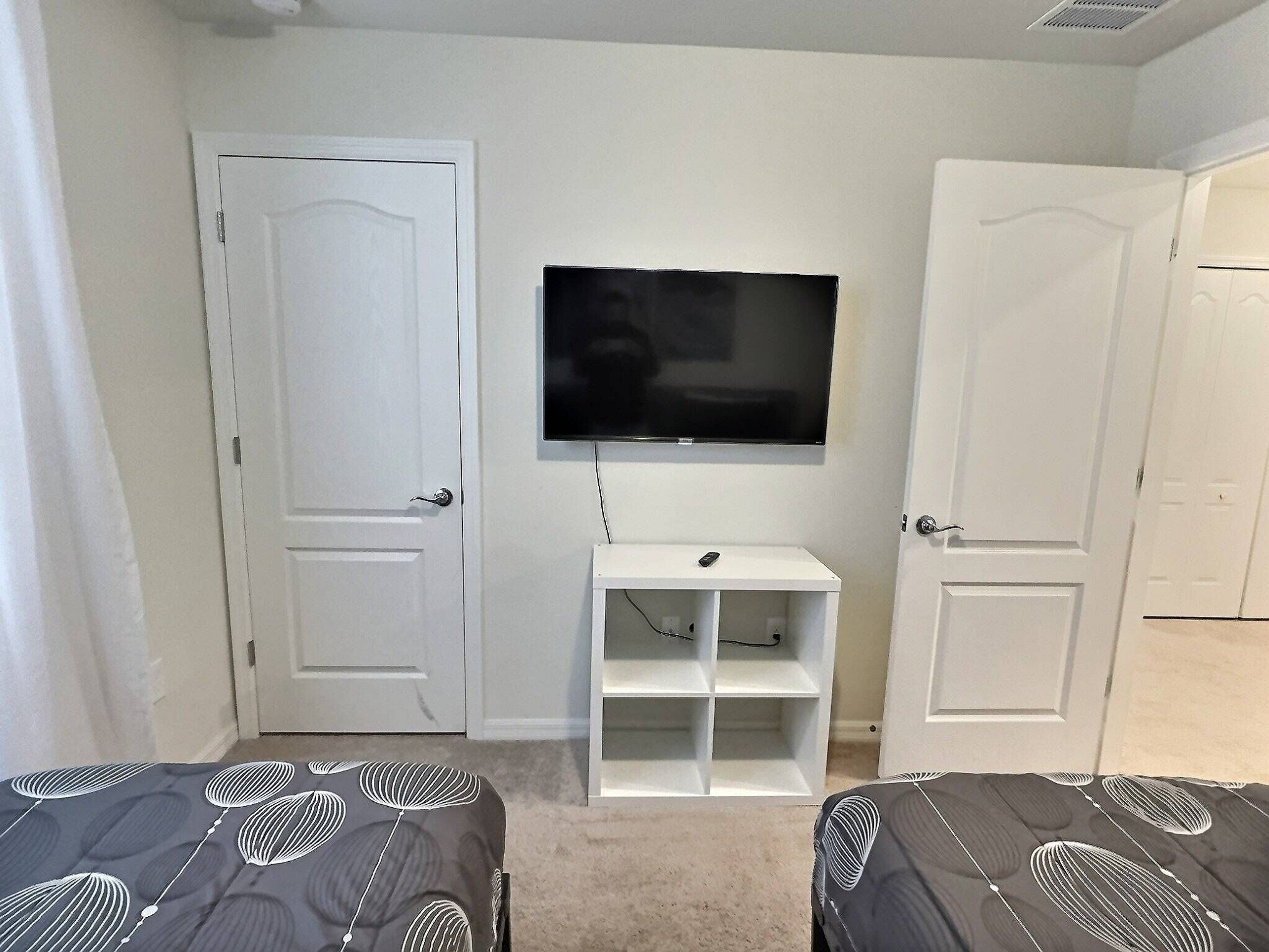 Ein Zimmer mit zwei Betten, einem an der Wand befestigten Fernseher und einem kleinen, weißen Regal.