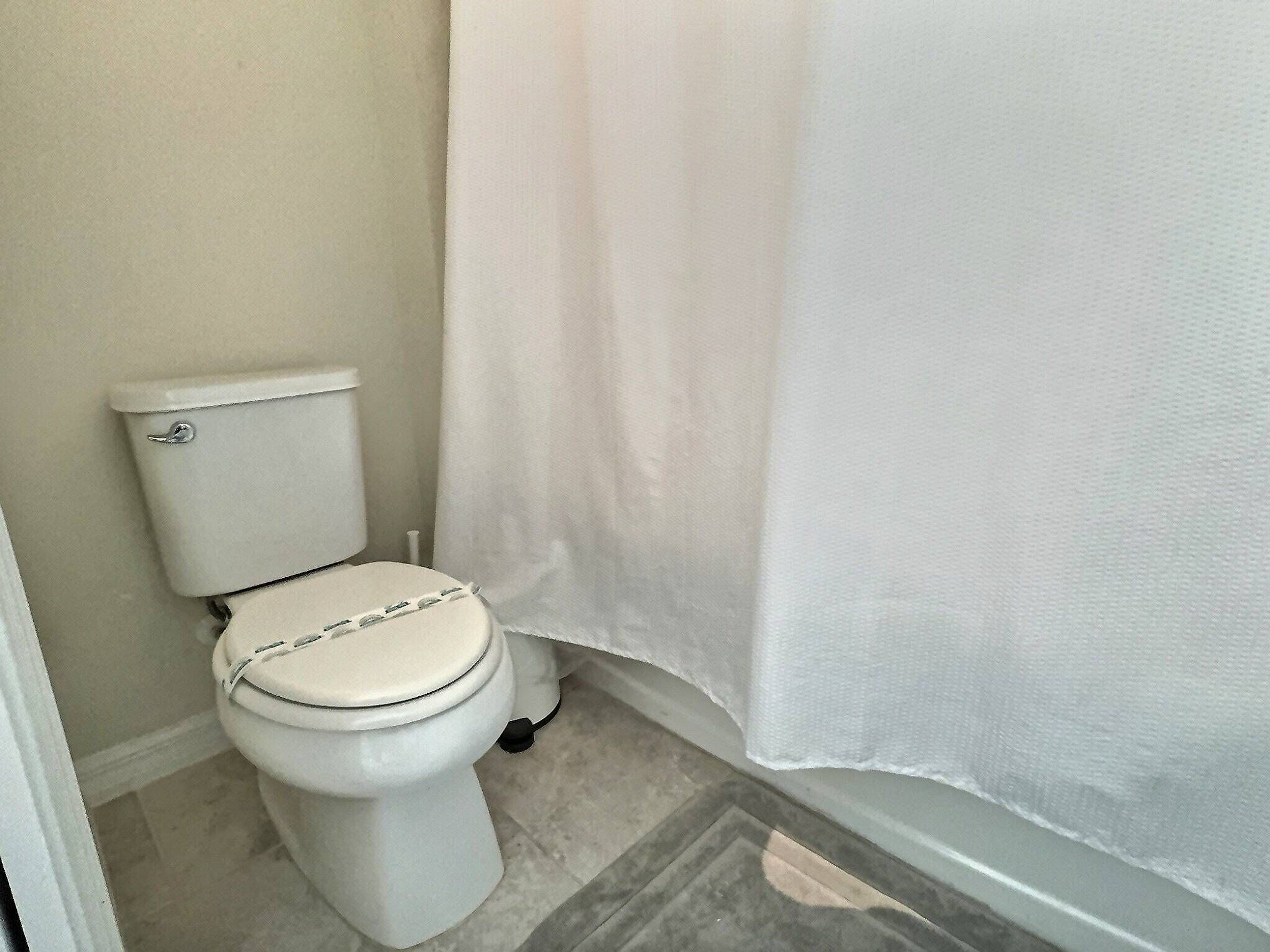 A small, white toilet with a closed lid, a white shower curtain, and a tiled floor.
