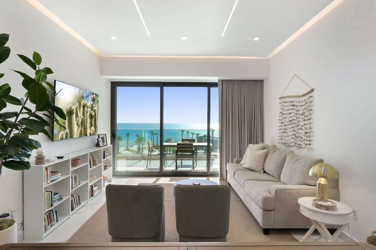 A modern living room with a sofa, two armchairs, a coffee table, a bookshelf, and a large window offering a view of the ocean.