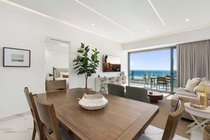 A modern living room with a dining area, a large window with a view of the sea, and a balcony with outdoor seating.
