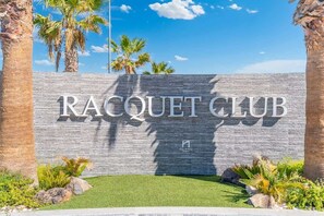 Schild für 'Racquet Club' vor einer Mauer aus Stein, Palmen und blauem Himmel.