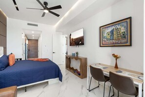 A modern bedroom with a bed, a desk, and a chair. There is a ceiling fan, a painting on the wall, and a door leading to another room.