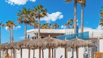 A beach with palm trees, sun loungers, and a building with a modern design.