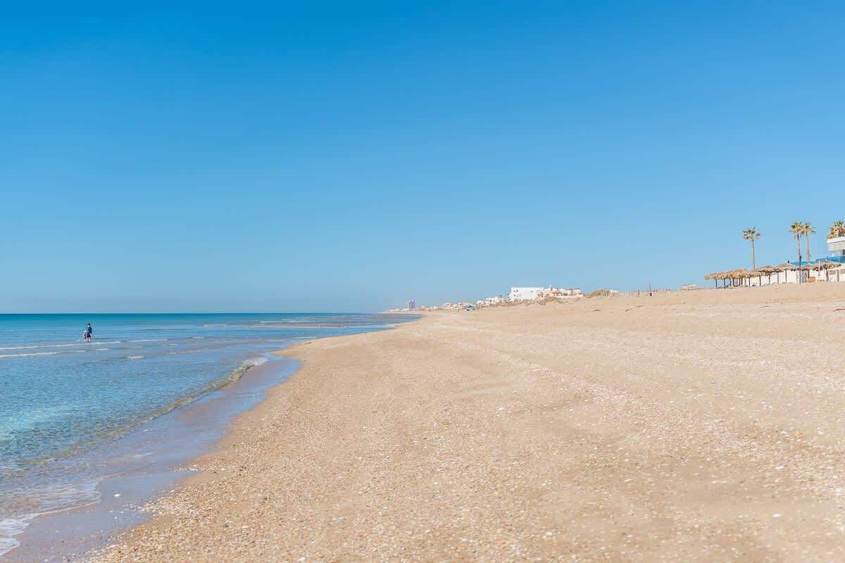 Une plage avec un ciel dégagé, une eau calme et quelques bâtiments visibles au loin.