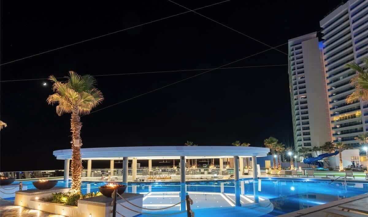Un espace piscine aménagé pour la nuit, avec un bar et des fauteuils confortables, entouré de palmiers et bordé d’un grand immeuble.