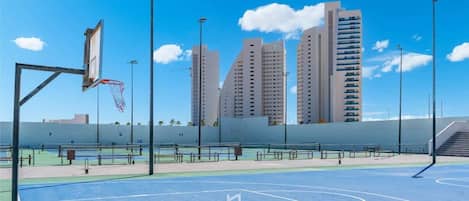 An outdoor basketball court with a basketball hoop, surrounded by tall buildings and clear skies.