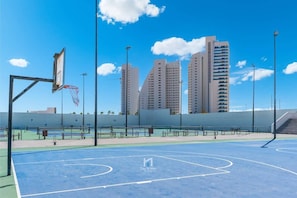Ein Basketballplatz im Freien mit Basketballkorb, umgeben von hohen Gebäuden und blauem Himmel.