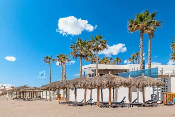Una playa con sombrillas de paja, palmeras y un edificio moderno con piscina.