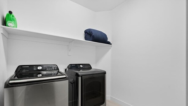 Ett tvättrum med en tvättmaskin och torktumlare från Samsung, en hylla med en grön flaska och en blå kudde.