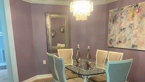Zona pranzo con tavolo in vetro circondato da quattro sedie, un lampadario, un grande quadro astratto e uno specchio sulla parete.